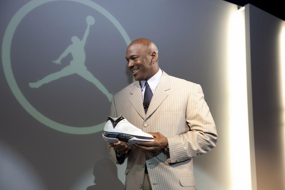Michael Jordan next to a Air Jordan Logo on a wall with a sneaker in his hand.